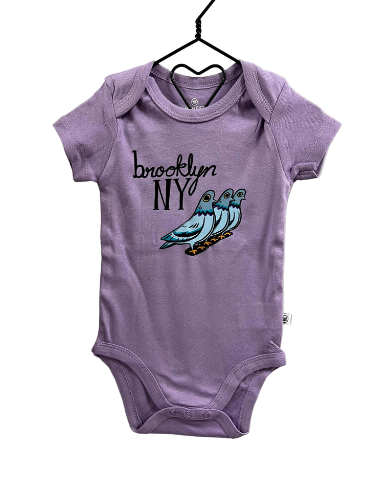 A lavender baby bodysuit hanging on a black hanger against a light gray wall. The bodysuit features a graphic print of three blue and teal pigeons with the text &