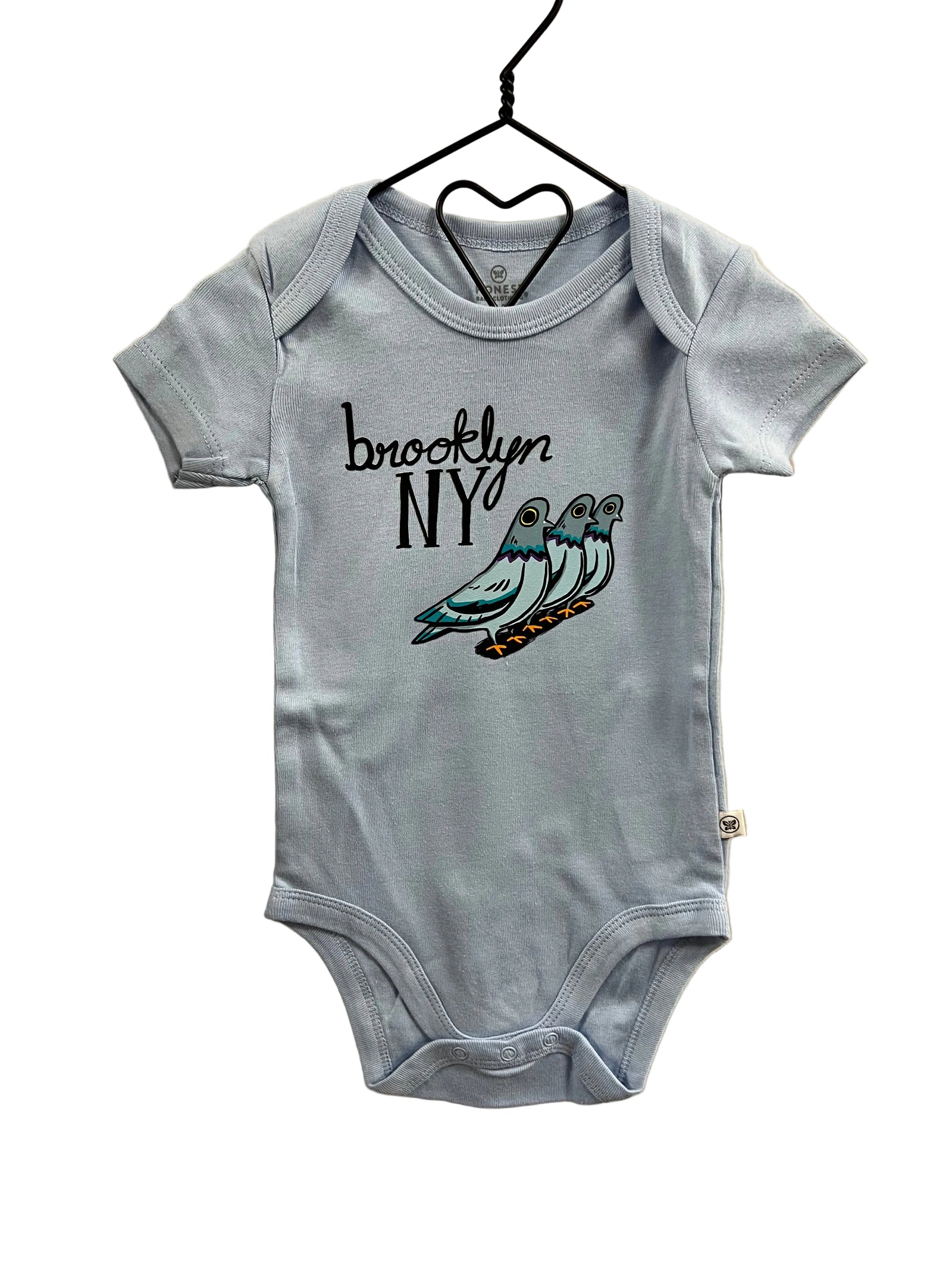 A light blue baby bodysuit hanging on a black hanger against a beige wall. The bodysuit features a graphic print on the front with the text 'brooklyn NY' in black script and an illustration of three pigeons in blue, gray, and orange.