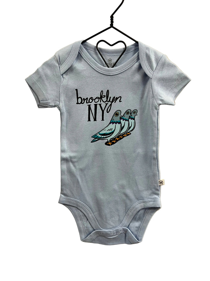 A light blue baby bodysuit hanging on a black hanger against a beige wall. The bodysuit features a graphic print on the front with the text &