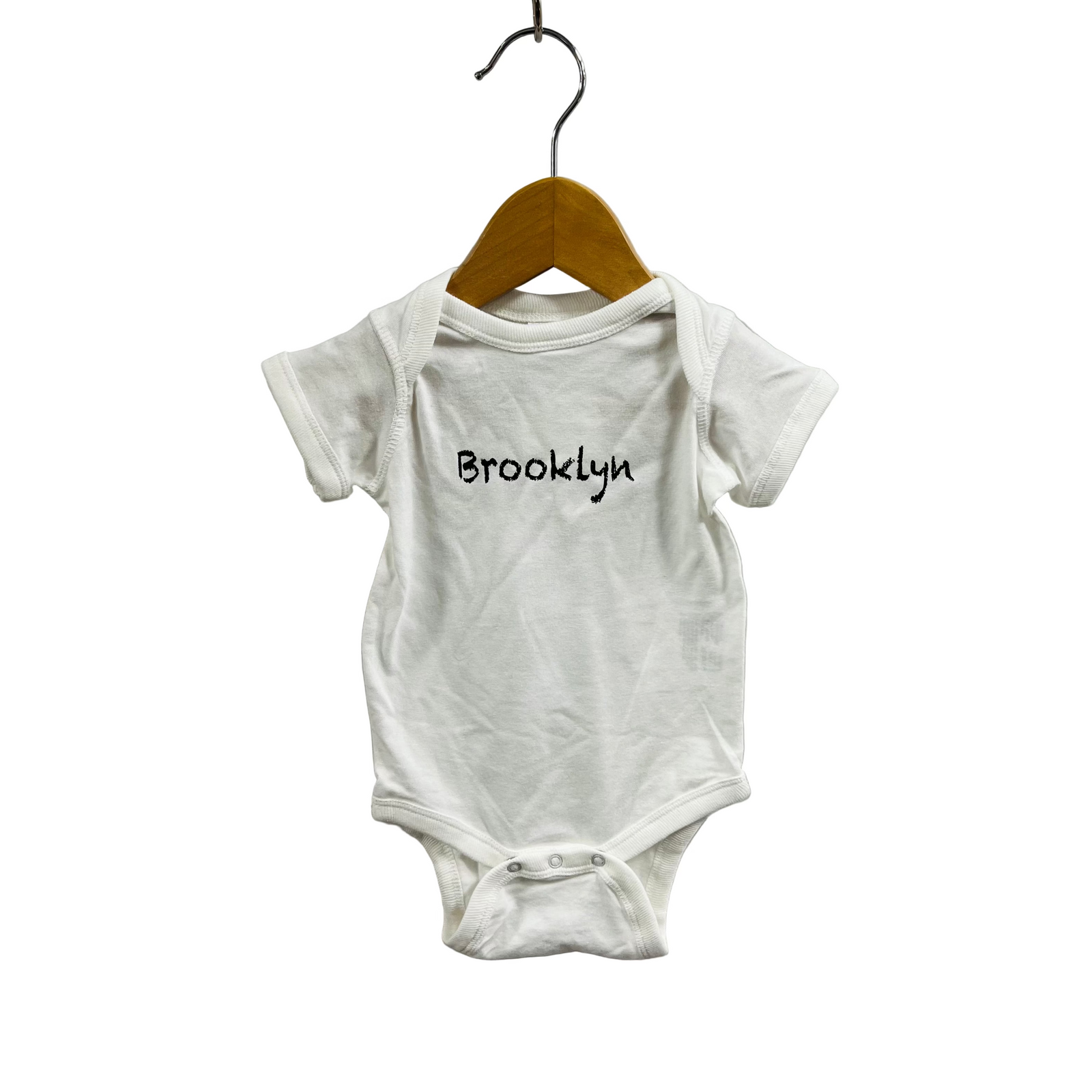 White baby onesie with 'Brooklyn' printed on it, hanging on a wooden hanger against a white background.