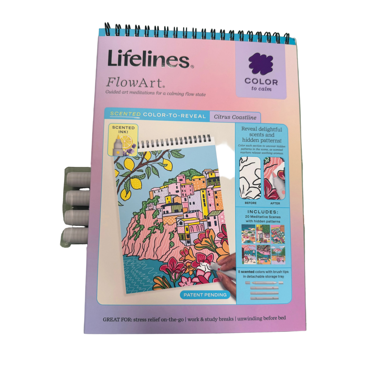 A Lifelines FlowArt color-to-reveal meditation pad titled 'Citrus Coastline' lies on a green surface next to several gray markers. The pad has a pink and lavender cover with a spiral binding at the top and a colorful illustrated cover showing a coastal village scene.