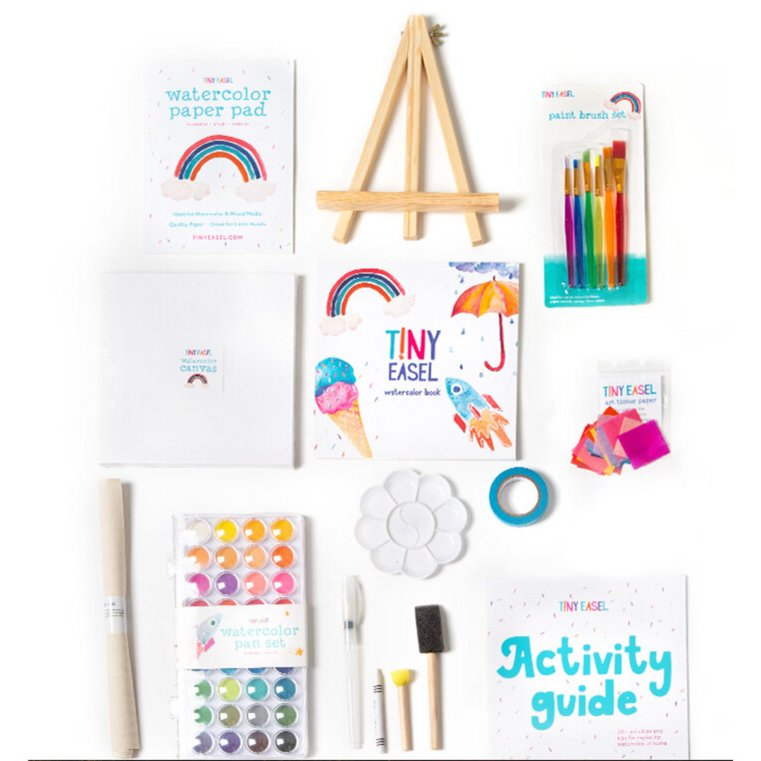An array of art supplies including watercolor paints, brushes, pencils, paper, and an easel arranged on a white background.