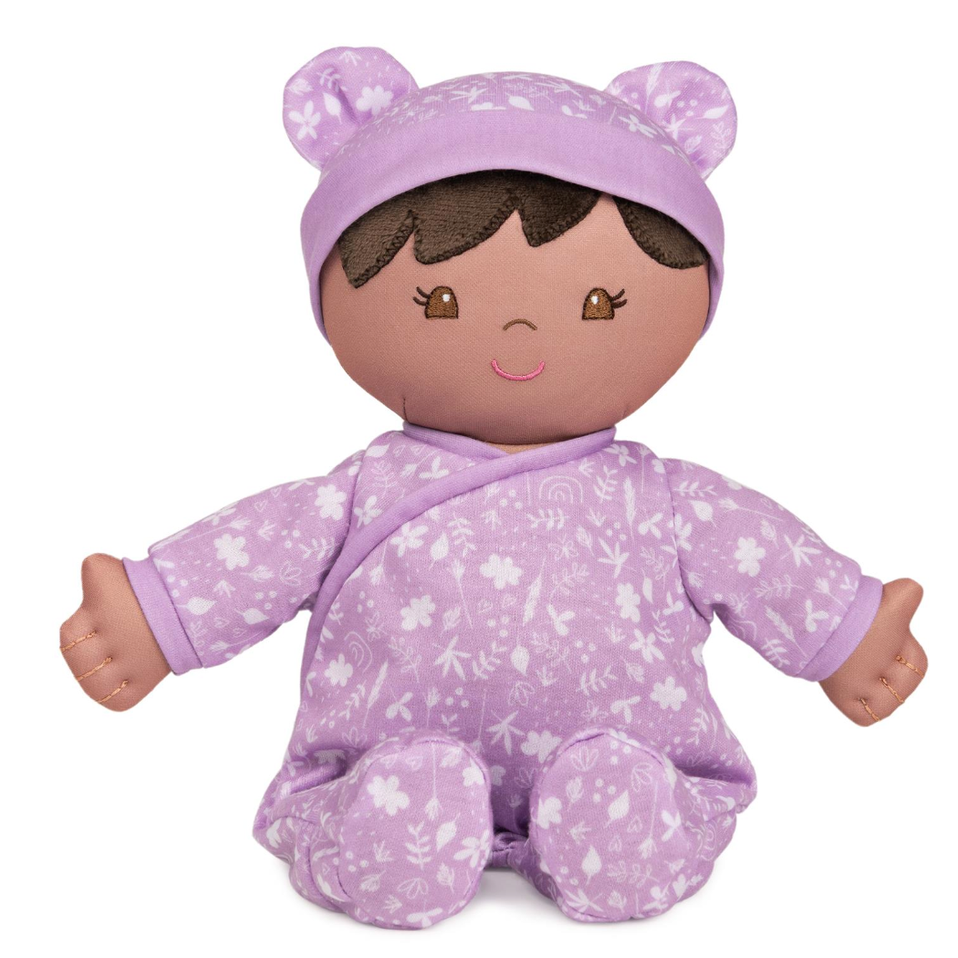 A plush baby doll with a lavender floral-patterned onesie and a matching hat with teddy bear ears. The doll has a brown body and is made of recycled plush material.